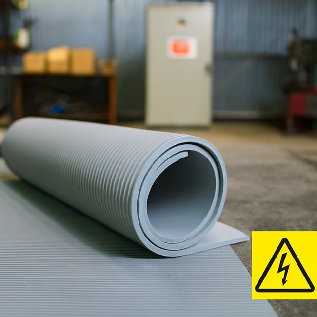 Rolled-up grey electrical insulation mat with ribbed surface, unrolled on the floor in an industrial workshop with high voltage warning symbol.